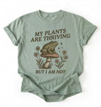 Flat lay of a bay sweatshirt with frog on mushroom illustration and text my plants are thriving but I am not on a white background