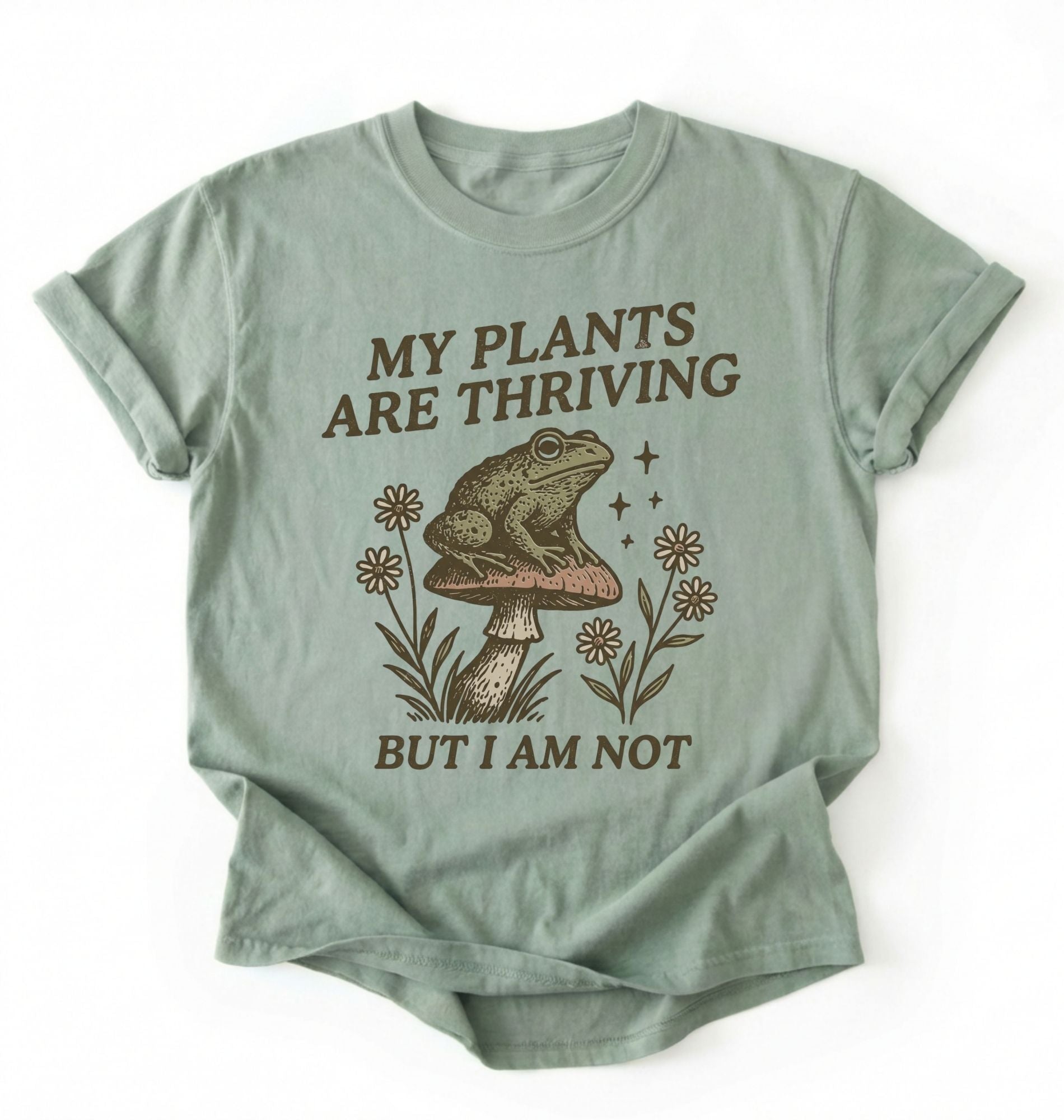 Flat lay of a bay sweatshirt with frog on mushroom illustration and text my plants are thriving but I am not on a white background