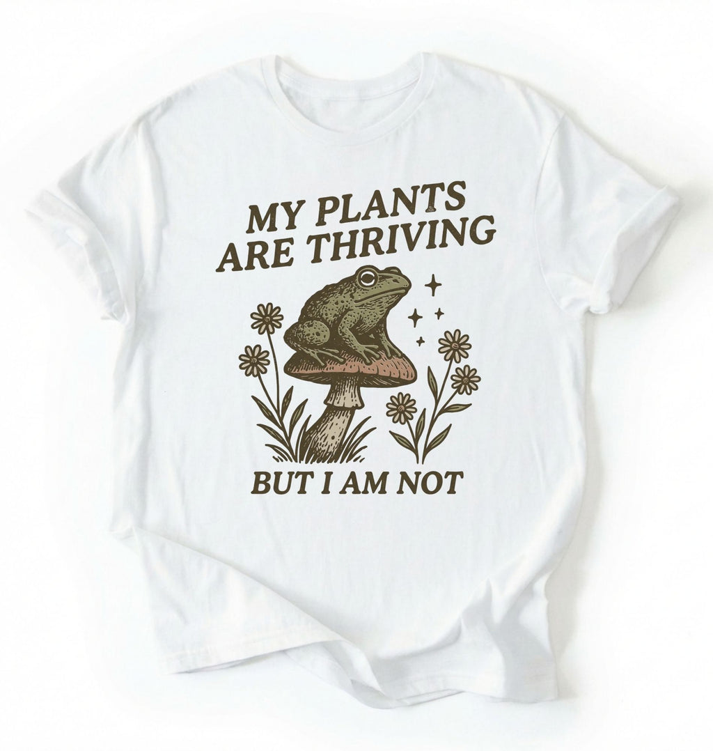 Flat lay of a white Comfort Colors sweatshirt with frog on mushroom illustration and text my plants are thriving but I am not on a white background 
