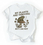 Flat lay of a white Comfort Colors sweatshirt with frog on mushroom illustration and text my plants are thriving but I am not on a white background 