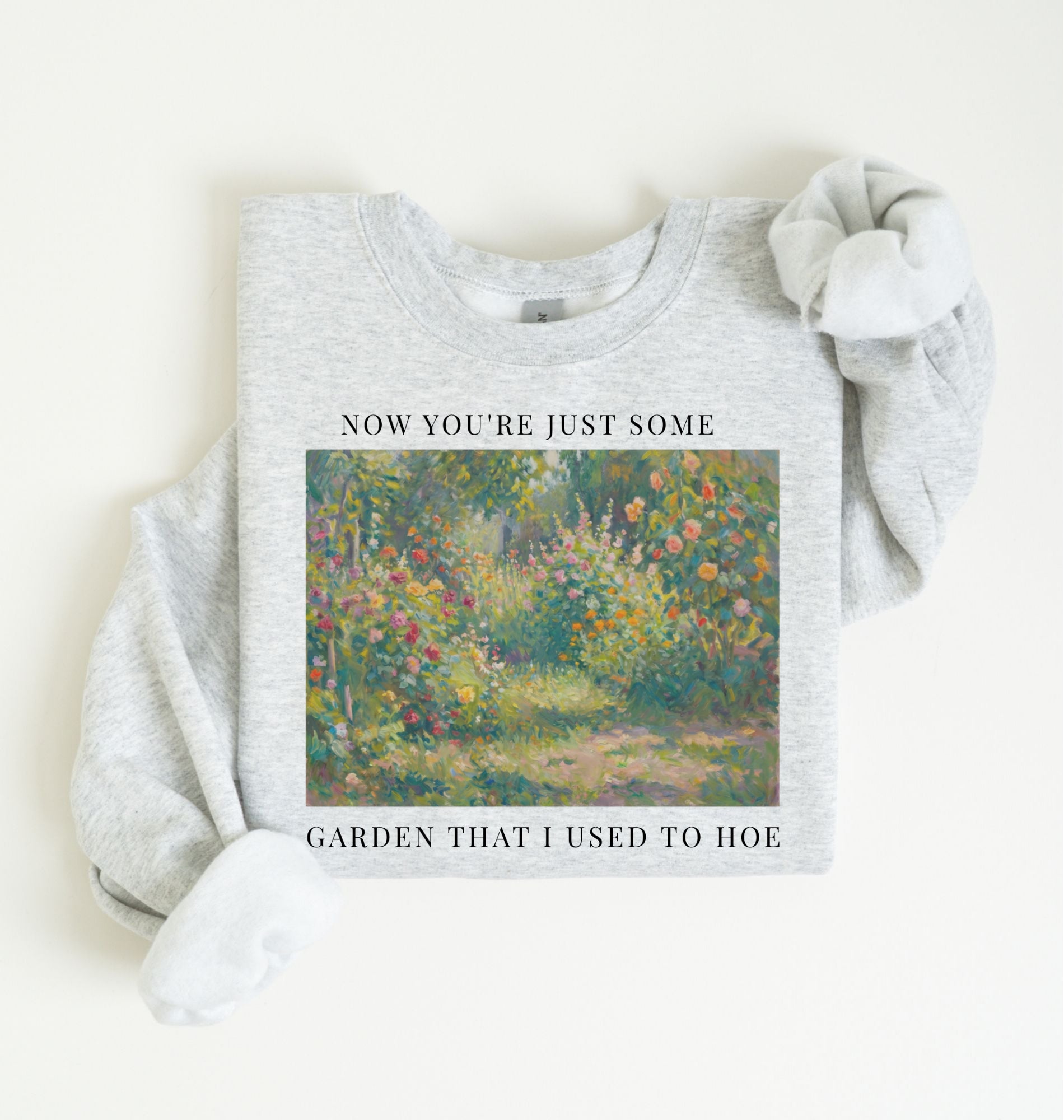 Flat lay of a ash crewneck sweatshirt on a white background, featuring an impressionist floral garden artwork and white text reading “Now you’re just some garden that I used to hoe.”
