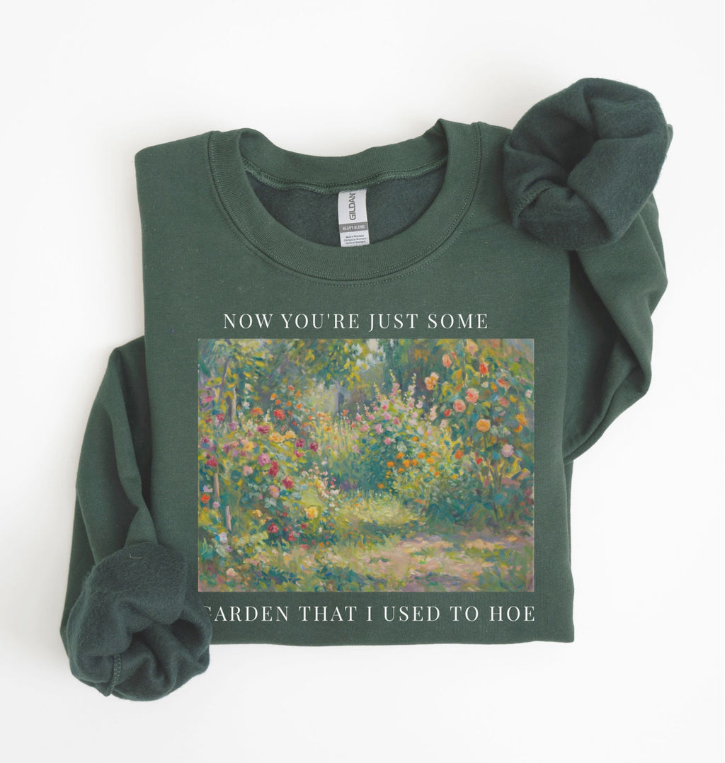 Flat lay of a forest green crewneck sweatshirt on a white background, featuring an impressionist floral garden artwork and white text reading “Now you’re just some garden that I used to hoe.