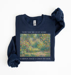Flat lay of a navy blue crewneck sweatshirt on a white background, featuring an impressionist floral garden artwork and white text reading “Now you’re just some garden that I used to hoe.