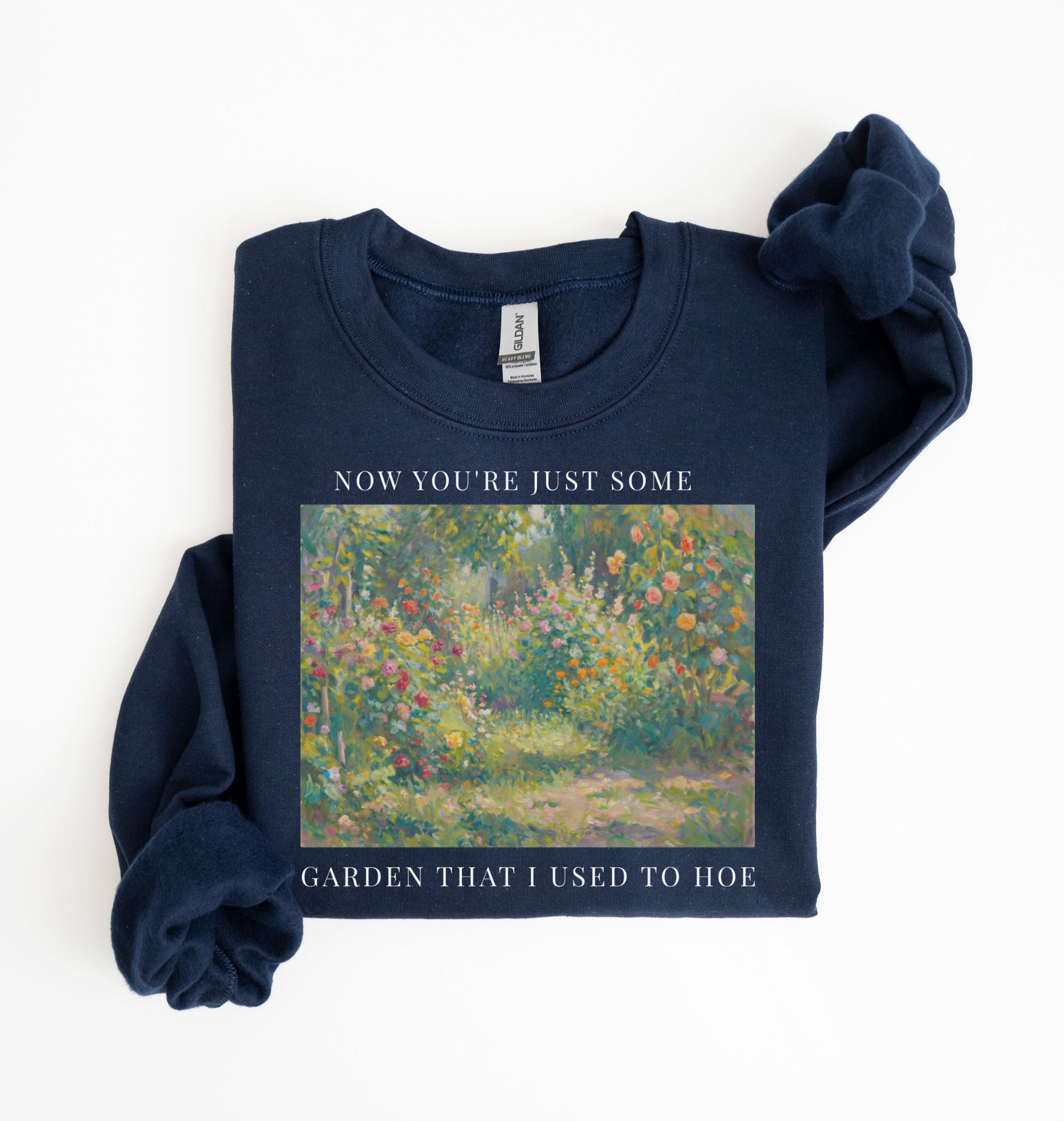 Flat lay of a navy blue crewneck sweatshirt on a white background, featuring an impressionist floral garden artwork and white text reading “Now you’re just some garden that I used to hoe.