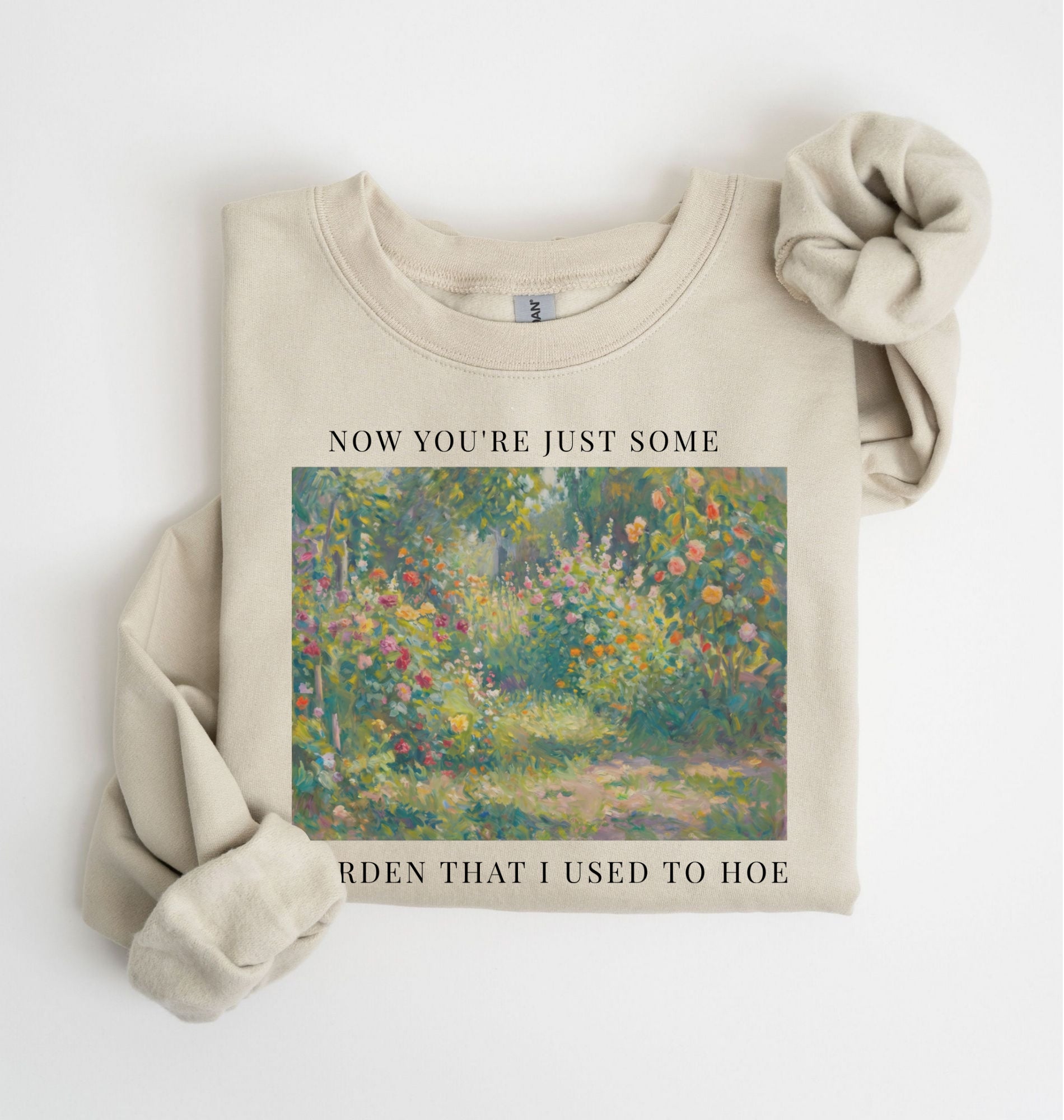 Flat lay of a sand crewneck sweatshirt on a white background, featuring an impressionist floral garden artwork and white text reading “Now you’re just some garden that I used to hoe.”