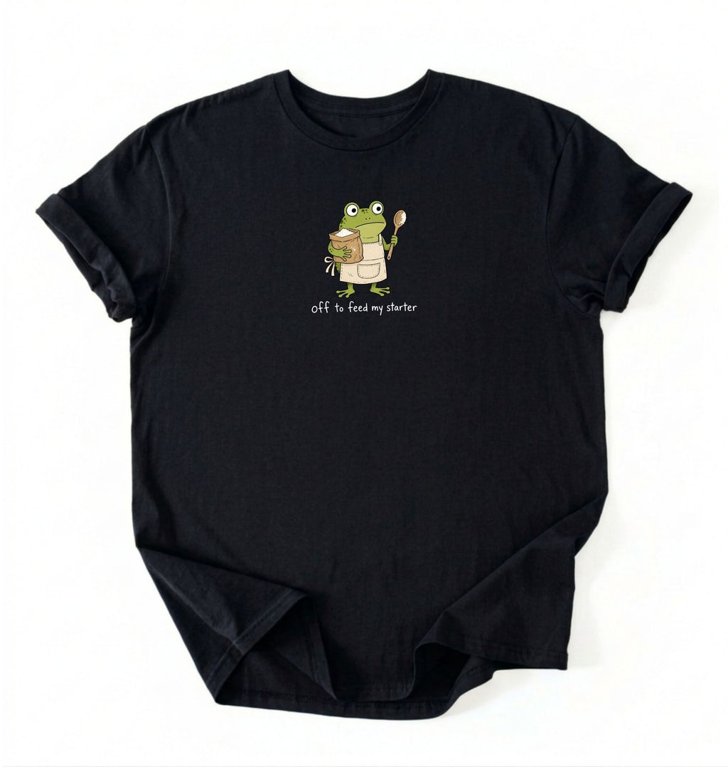 A black short-sleeve crewneck t-shirt laid flat on a white background, featuring a cartoon graphic of a green frog holding a spoon and a bowl, with the text 'Off to feed my starter' printed below.