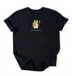 A black short-sleeve crewneck t-shirt laid flat on a white background, featuring a cartoon graphic of a green frog holding a spoon and a bowl, with the text 'Off to feed my starter' printed below.