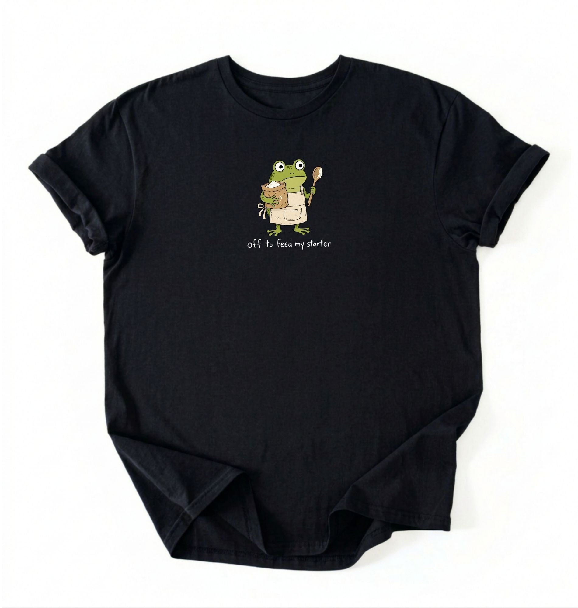 A black short-sleeve crewneck t-shirt laid flat on a white background, featuring a cartoon graphic of a green frog holding a spoon and a bowl, with the text 'Off to feed my starter' printed below.