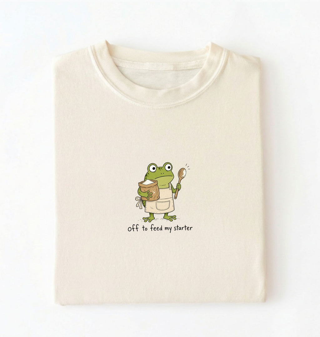 A folded cream-colored t-shirt with a cartoon graphic of a green frog wearing an apron and holding a spoon next to a jar, with the text 'off to feed my starter' printed below.