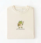 A folded cream-colored t-shirt with a cartoon graphic of a green frog wearing an apron and holding a spoon next to a jar, with the text 'off to feed my starter' printed below.