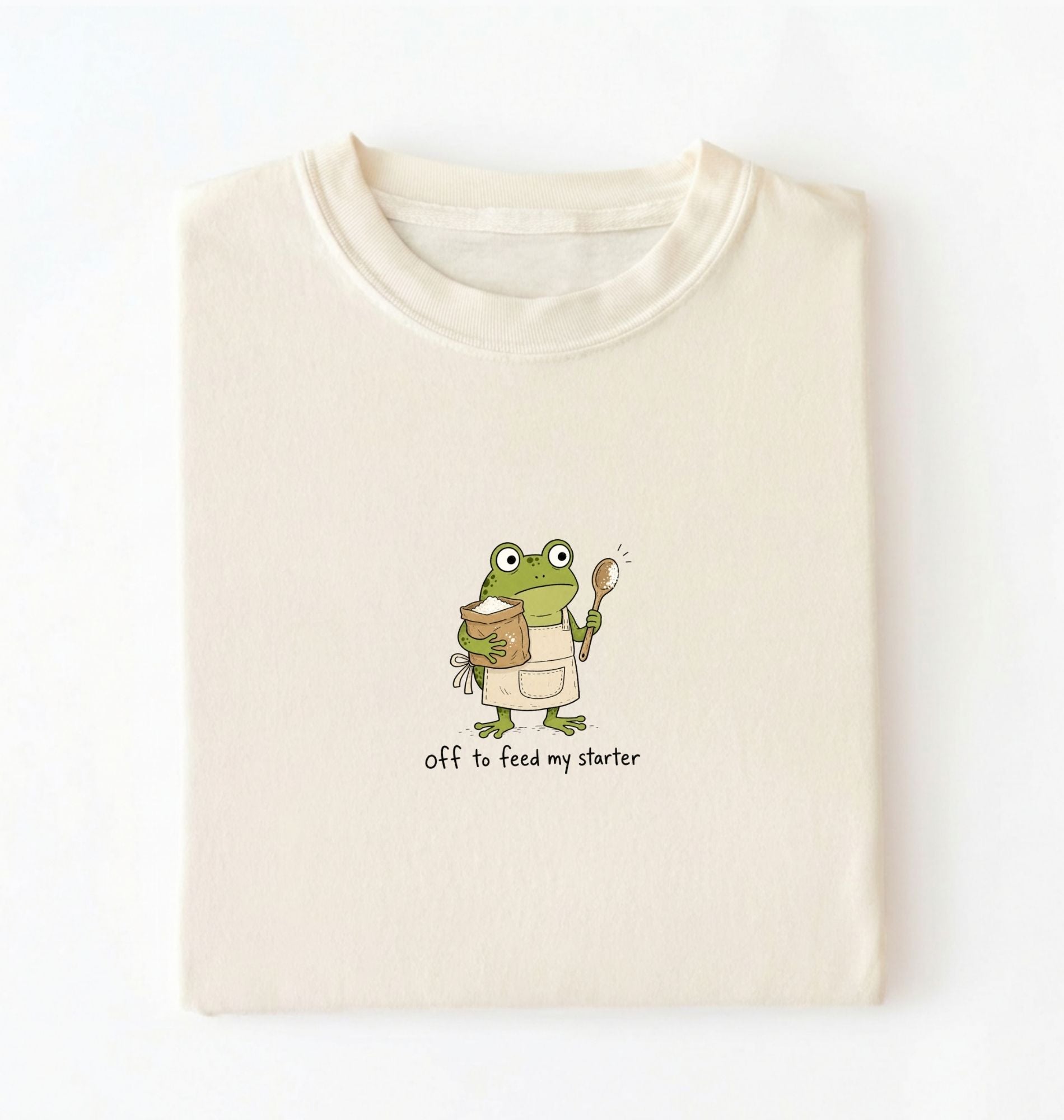 A folded cream-colored t-shirt with a cartoon graphic of a green frog wearing an apron and holding a spoon next to a jar, with the text 'off to feed my starter' printed below.