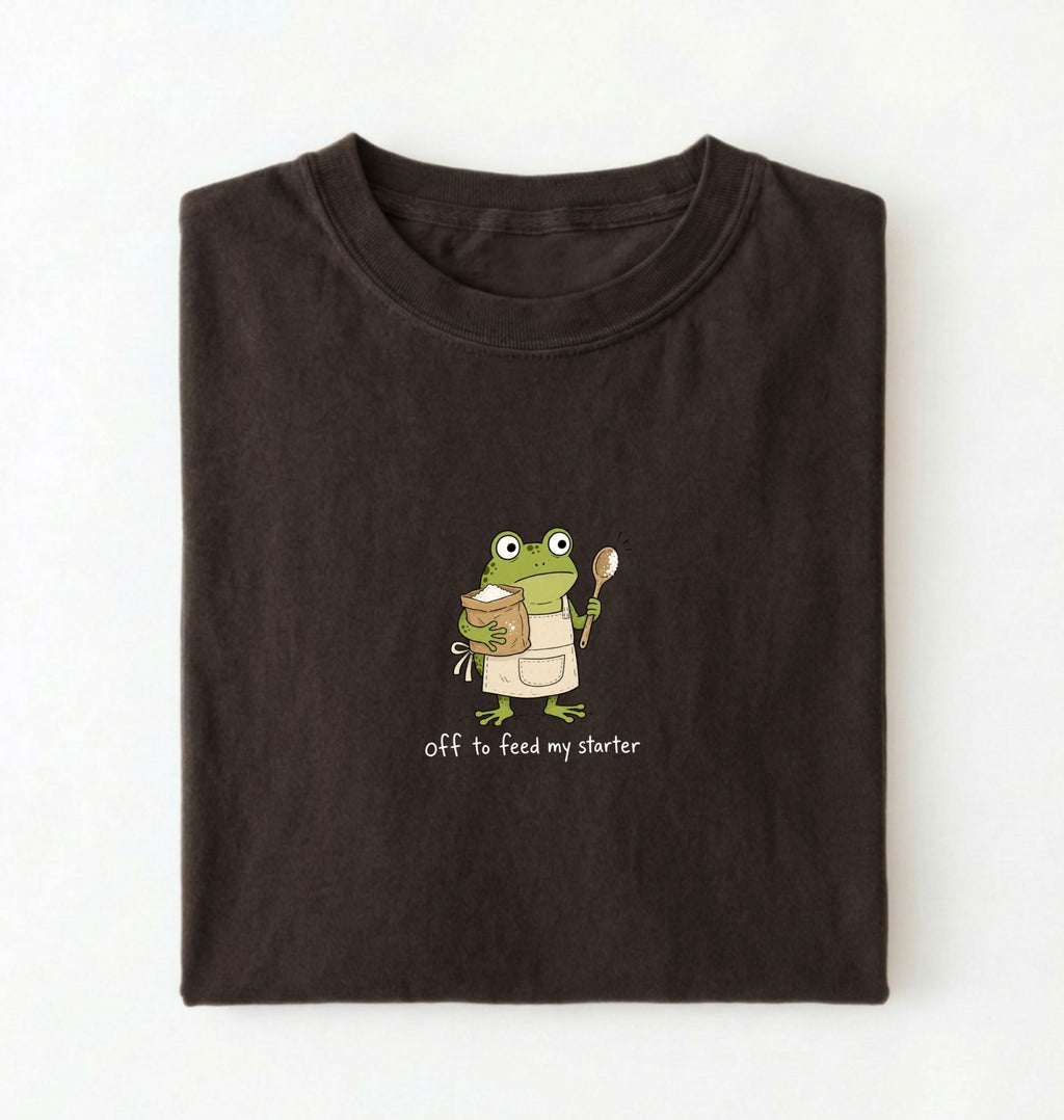 A dark brown folded t-shirt featuring a cartoon green frog wearing a beige apron and holding a spoon next to a mug, with the text 'off to feed my starter' printed on a white background 