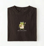 A dark brown folded t-shirt featuring a cartoon green frog wearing a beige apron and holding a spoon next to a mug, with the text 'off to feed my starter' printed on a white background 