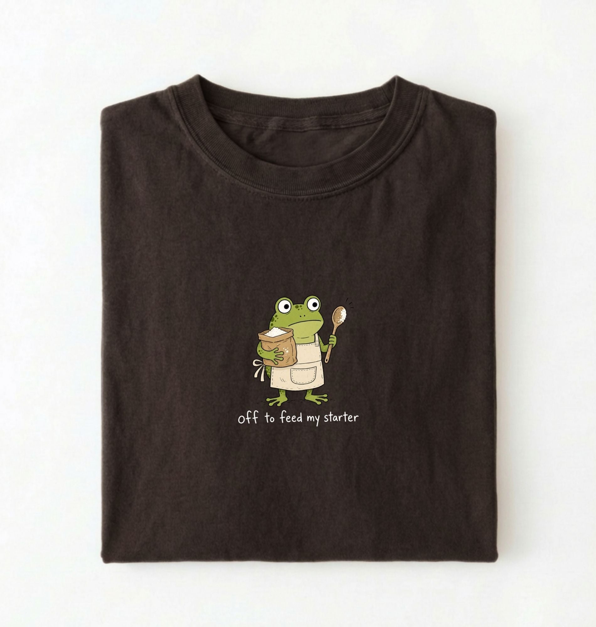A dark brown folded t-shirt featuring a cartoon green frog wearing a beige apron and holding a spoon next to a mug, with the text 'off to feed my starter' printed on a white background 