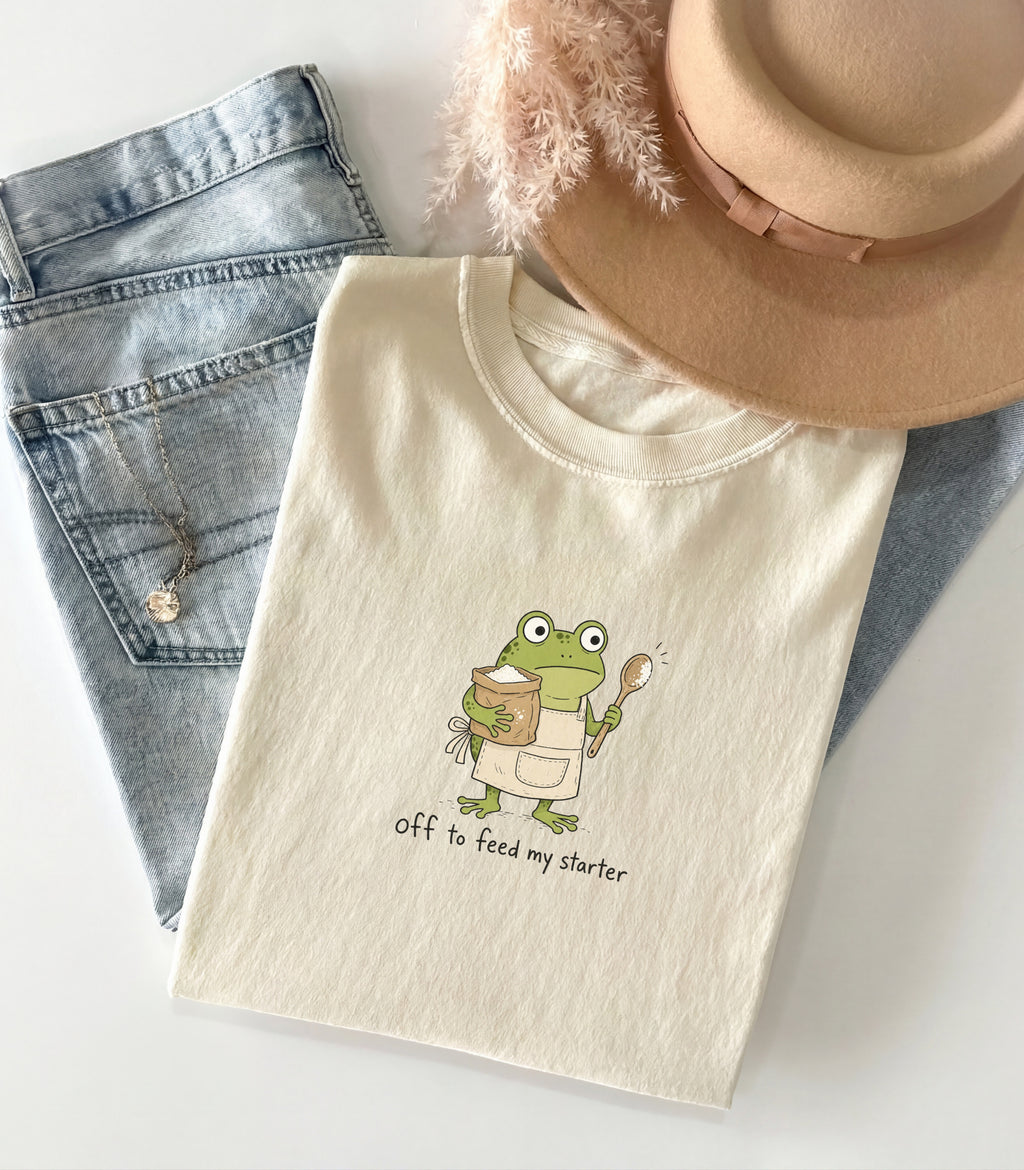 A folded cream-colored t-shirt with a cute cartoon frog graphic holding a spoon and a mug, with the text 'off to feed my starter' printed below. The t-shirt is styled in a flat-lay arrangement with light-wash blue jeans, a beige fedora hat, and a fluffy beige feather on a white background.