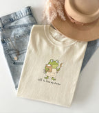 A folded cream-colored t-shirt with a cute cartoon frog graphic holding a spoon and a mug, with the text 'off to feed my starter' printed below. The t-shirt is styled in a flat-lay arrangement with light-wash blue jeans, a beige fedora hat, and a fluffy beige feather on a white background.