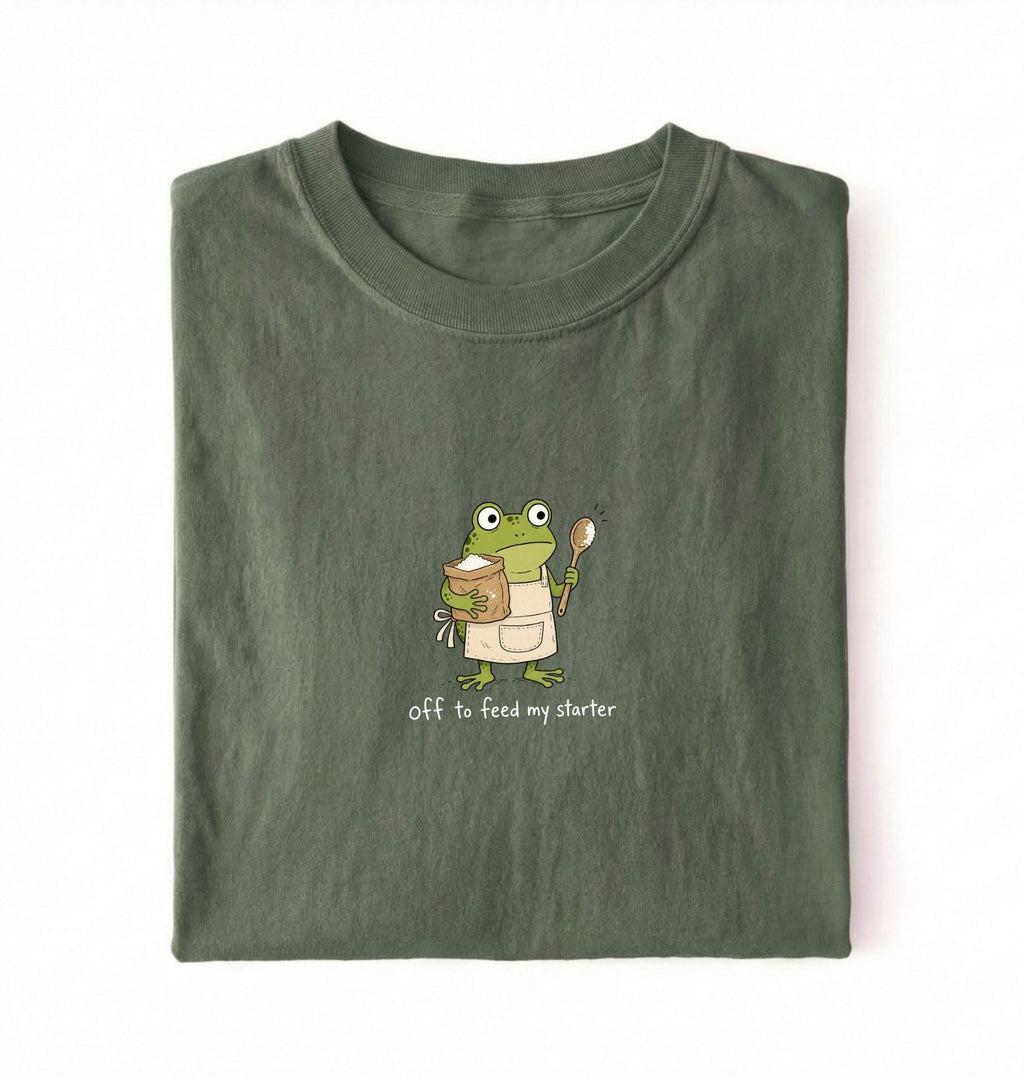 Flat lay of a green sourdough shirt featuring a frog baker and the phrase Off to Feed My Starter on a white background