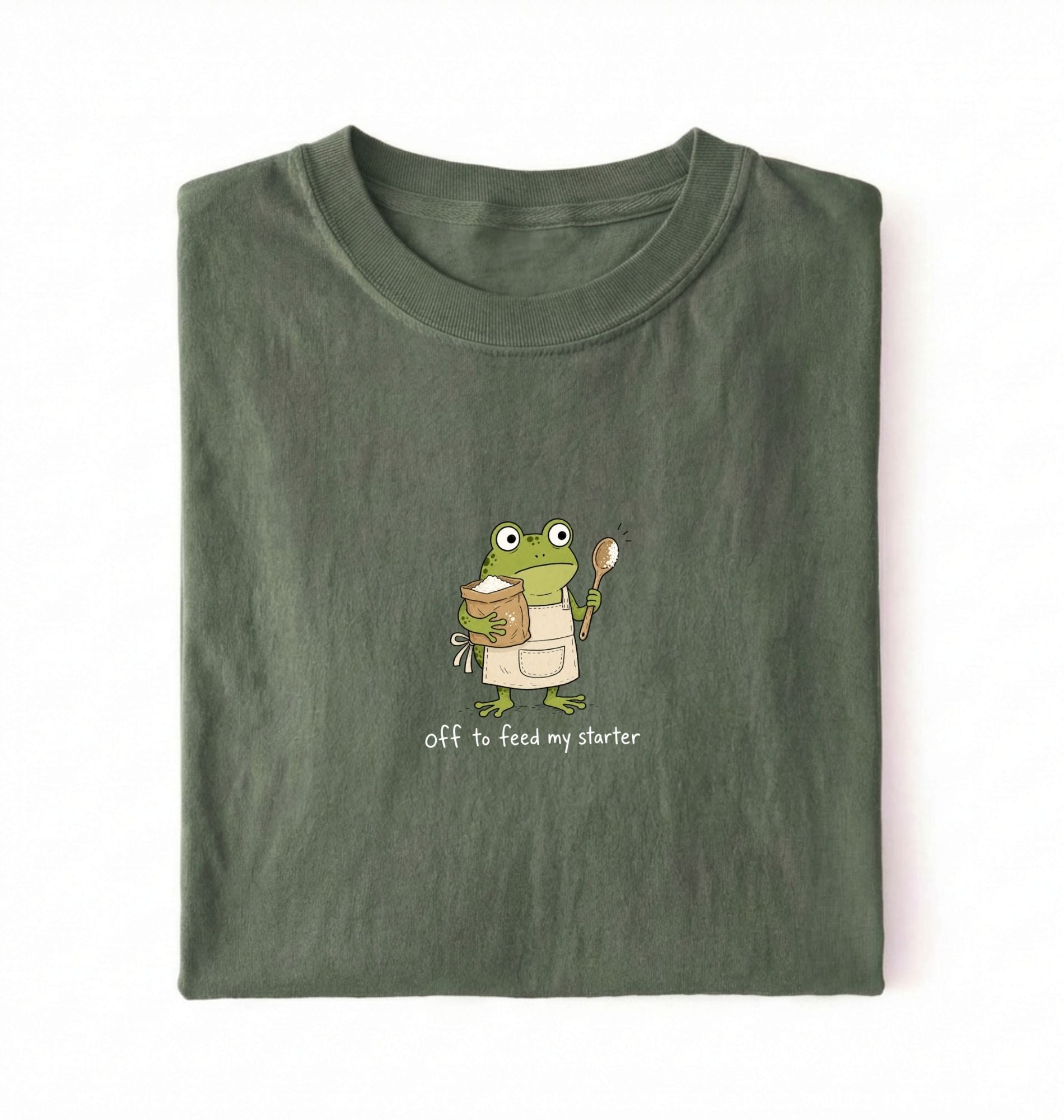 Flat lay of a green sourdough shirt featuring a frog baker and the phrase Off to Feed My Starter on a white background