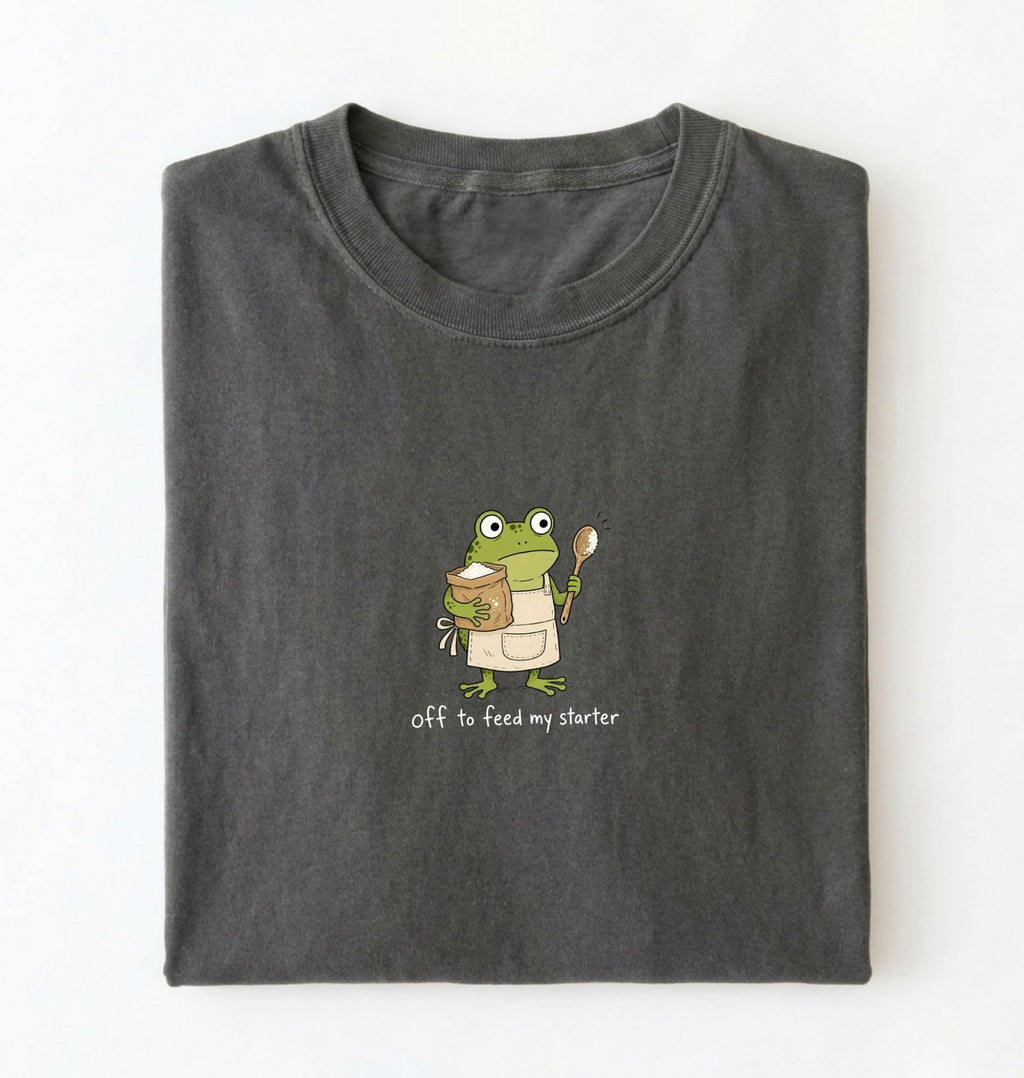 A folded dark gray t-shirt on a white background, featuring a cartoon green frog wearing an apron and holding a spoon, with the text 'off to feed my starter' printed below.