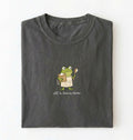 A folded dark gray t-shirt on a white background, featuring a cartoon green frog wearing an apron and holding a spoon, with the text 'off to feed my starter' printed below.