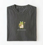 A folded dark gray t-shirt on a white background, featuring a cartoon green frog wearing an apron and holding a spoon, with the text 'off to feed my starter' printed below.