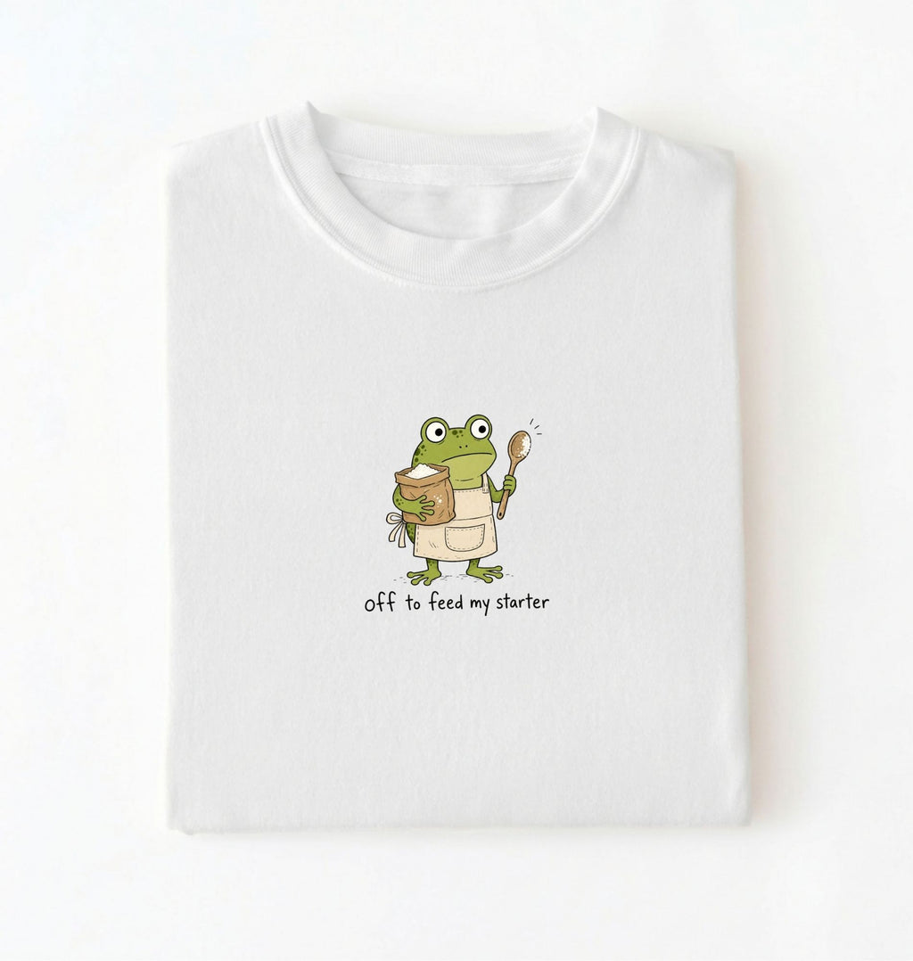 A folded white t-shirt with a cartoon graphic of a green frog wearing an apron and holding a spoon next to a jar, with the text 'off to feed my starter' on a white background