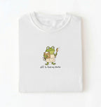 A folded white t-shirt with a cartoon graphic of a green frog wearing an apron and holding a spoon next to a jar, with the text 'off to feed my starter' on a white background