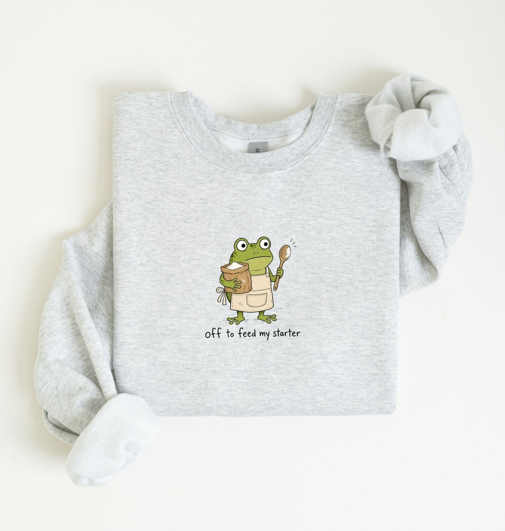 A light heather gray long-sleeve sweatshirt laid flat on a cream background, featuring a cartoon green frog wearing an apron and holding a spoon, with the text 'off to feed my starter' printed below.