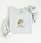 A light heather gray long-sleeve sweatshirt laid flat on a cream background, featuring a cartoon green frog wearing an apron and holding a spoon, with the text 'off to feed my starter' printed below.
