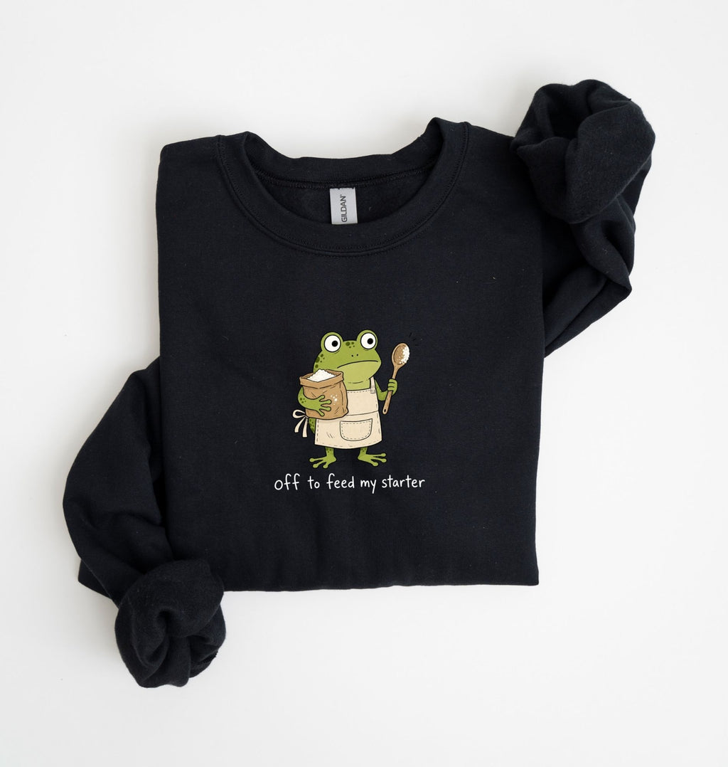 A black long-sleeve sweatshirt with a graphic of a green cartoon frog wearing an apron and holding a mug, with the text 'off to feed my starter' printed below. The sweatshirt is laid flat on a white background with one sleeve rolled up.