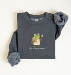 A folded dark heather gray sweatshirt with rolled sleeves, featuring a cartoon green frog holding a spoon and book, with the text 'off to feed my starter' printed below. The sweatshirt is laid flat on a light off-white background.