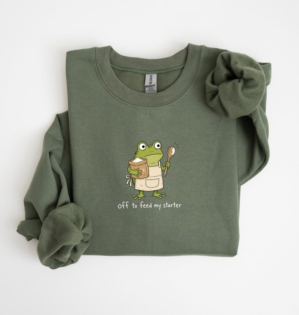 An olive green long-sleeve sweatshirt with a cartoon frog graphic holding a mug and spoon, with the text 'off to feed my starter' printed below. The sweatshirt is folded neatly on a white background.