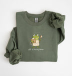 An olive green long-sleeve sweatshirt with a cartoon frog graphic holding a mug and spoon, with the text 'off to feed my starter' printed below. The sweatshirt is folded neatly on a white background.