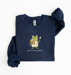 A navy blue long-sleeve sweatshirt with a cartoon frog graphic holding a spoon and a mug, with the text 'off to feed my starter' printed below. The sweatshirt is laid flat on a white background with one sleeve folded over.