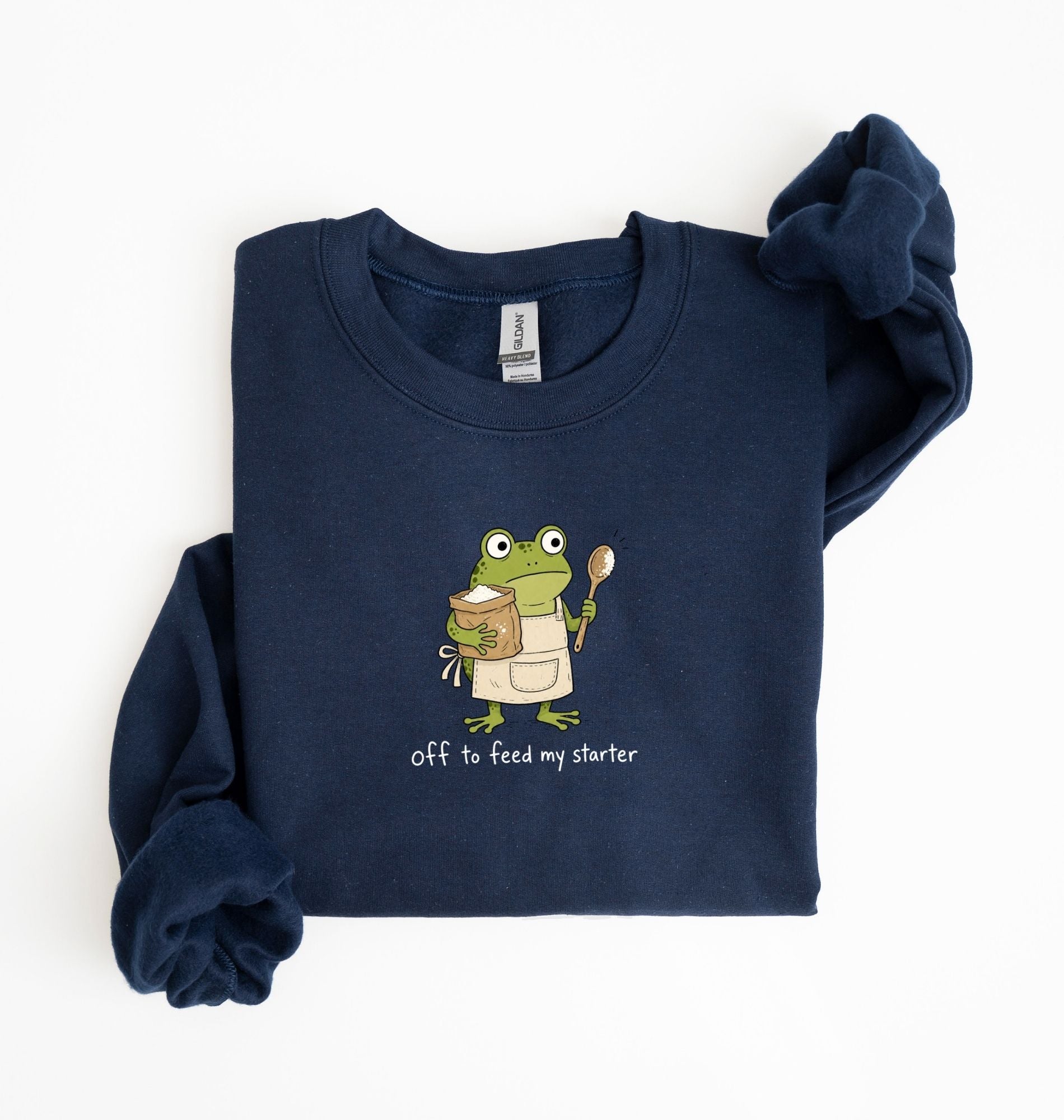 A navy blue long-sleeve sweatshirt with a cartoon frog graphic holding a spoon and a mug, with the text 'off to feed my starter' printed below. The sweatshirt is laid flat on a white background with one sleeve folded over.