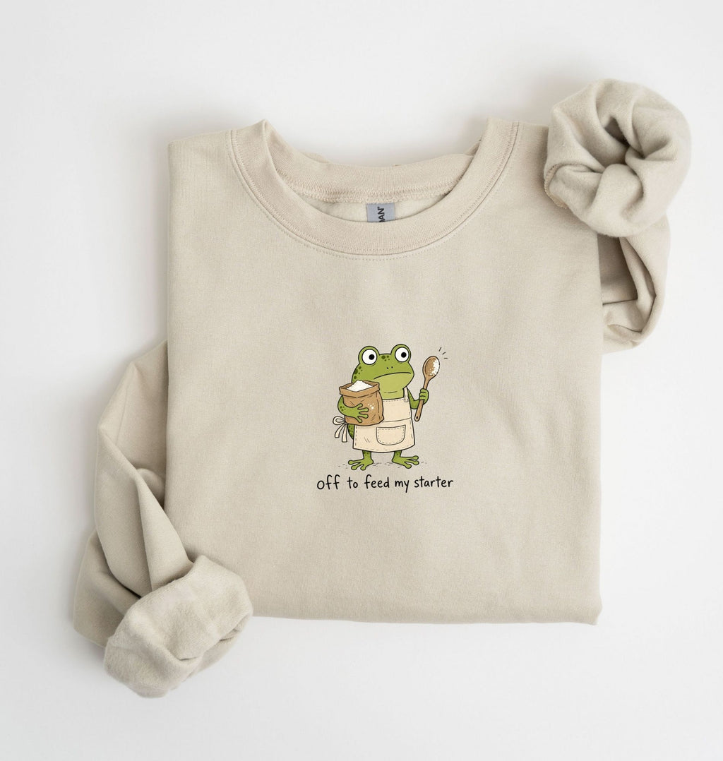 A beige crewneck sweatshirt with a graphic of a green cartoon frog holding a jar of peanut butter and a spoon, with the text 'Off to feed my starter' printed below. The sweatshirt is styled flat on a white background with one sleeve rolled up and the other cuff extended.