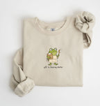 A beige crewneck sweatshirt with a graphic of a green cartoon frog holding a jar of peanut butter and a spoon, with the text 'Off to feed my starter' printed below. The sweatshirt is styled flat on a white background with one sleeve rolled up and the other cuff extended.