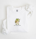A white long-sleeve sweatshirt laid flat on a white background, featuring a cartoon green frog holding a mug and spoon with the text 'Off to feed my starter' printed below.