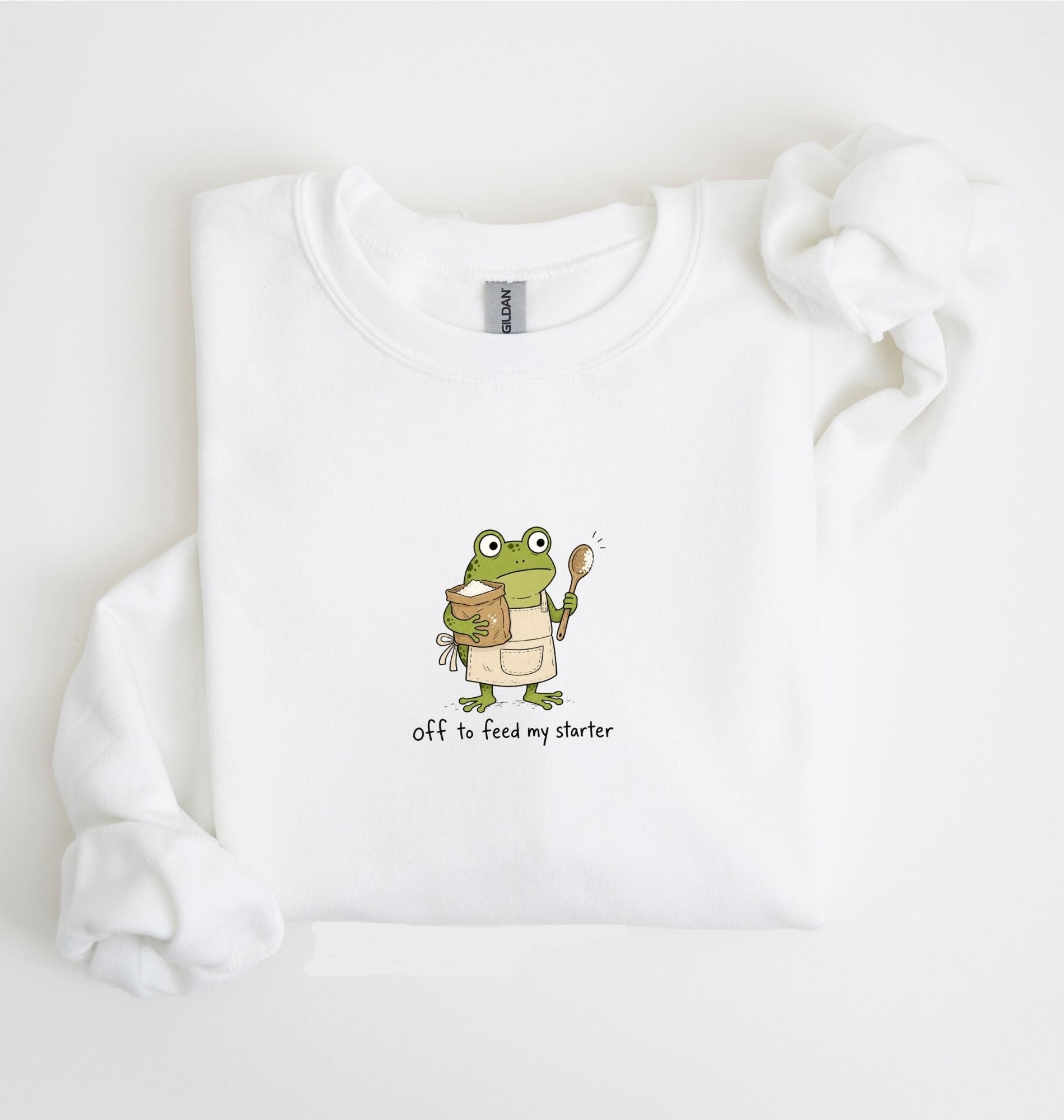 A white long-sleeve sweatshirt laid flat on a white background, featuring a cartoon green frog holding a mug and spoon with the text 'Off to feed my starter' printed below.
