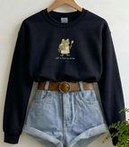 A black crewneck sweatshirt with a cartoon frog graphic holding a spoon and the text 'Off to feed my charmer' printed on the chest. The sweatshirt is styled on a wooden hanger with light-wash denim shorts and a brown belt against a plain white wall.