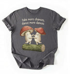 Flat lay of a Pepper charcoal t-shirt with two dancing mushrooms and the phrase take more chances dance more dances, whimsical cottagecore gift tee on a white background 