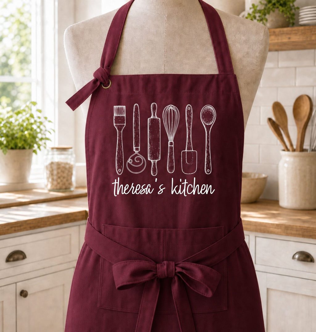 A burgundy apron with white line-art illustrations of kitchen utensils and the text 'theresa's kitchen' displayed on a mannequin torso in a bright kitchen setting.