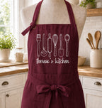 A burgundy apron with white line-art illustrations of kitchen utensils and the text 'theresa's kitchen' displayed on a mannequin torso in a bright kitchen setting.