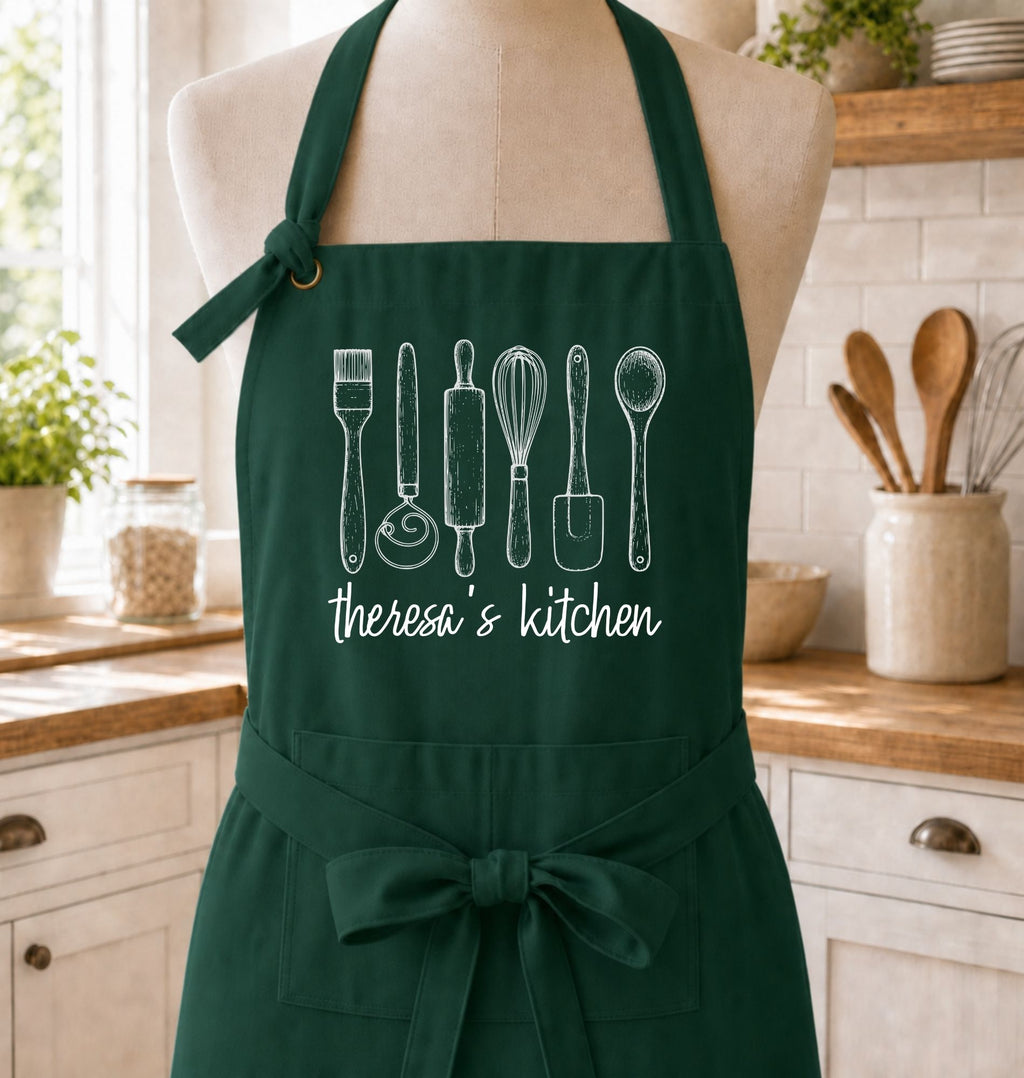 A dark green kitchen apron displayed on a mannequin torso in a bright kitchen setting. The apron features white line drawings of kitchen utensils and the text 'theresa's kitchen' printed on the front.