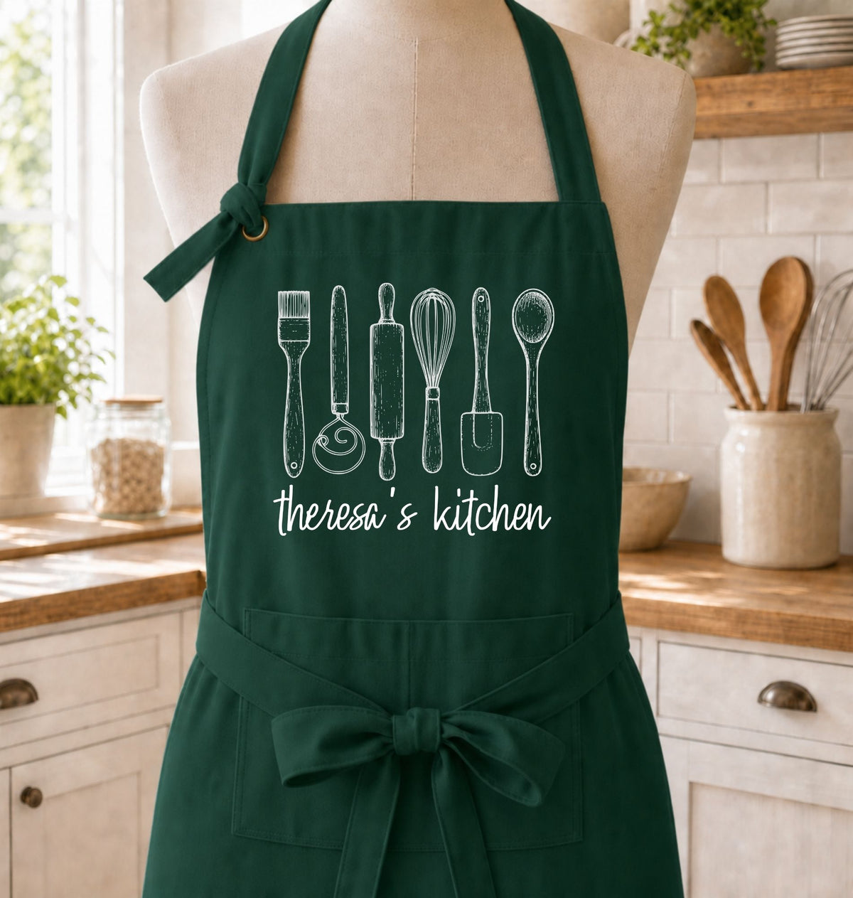 A dark green kitchen apron displayed on a mannequin torso in a bright kitchen setting. The apron features white line drawings of kitchen utensils and the text 'theresa's kitchen' printed on the front.