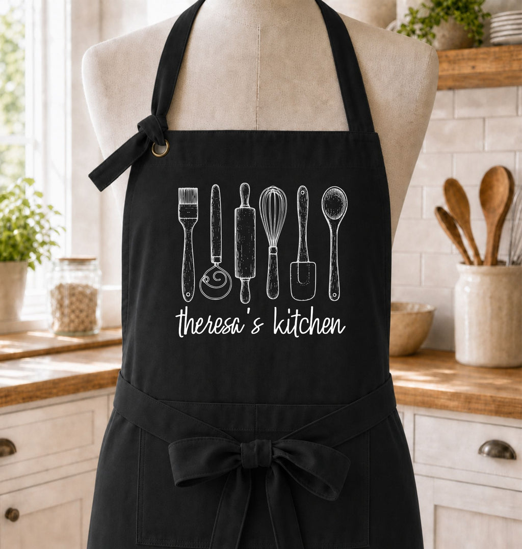 A black kitchen apron with white chalk-style drawings of cooking utensils and the text 'therosa's kitchen' displayed on a beige dress form in a bright kitchen setting.