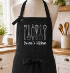 A black kitchen apron with white chalk-style drawings of cooking utensils and the text 'therosa's kitchen' displayed on a beige dress form in a bright kitchen setting.