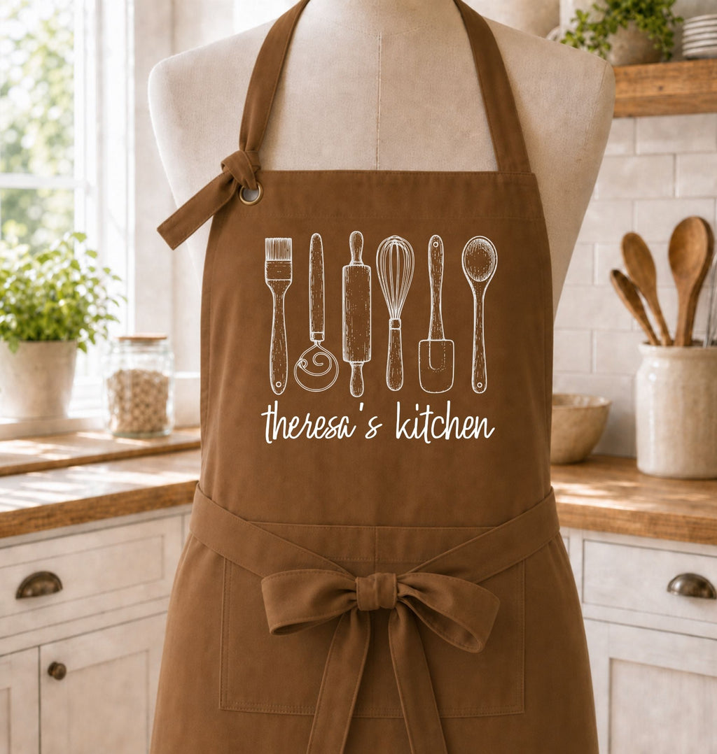 A brown kitchen apron with white line-art drawings of cooking utensils and the text 'theresa's kitchen' displayed on a mannequin torso in a bright, styled kitchen setting.