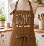 A brown kitchen apron with white line-art drawings of cooking utensils and the text 'theresa's kitchen' displayed on a mannequin torso in a bright, styled kitchen setting.