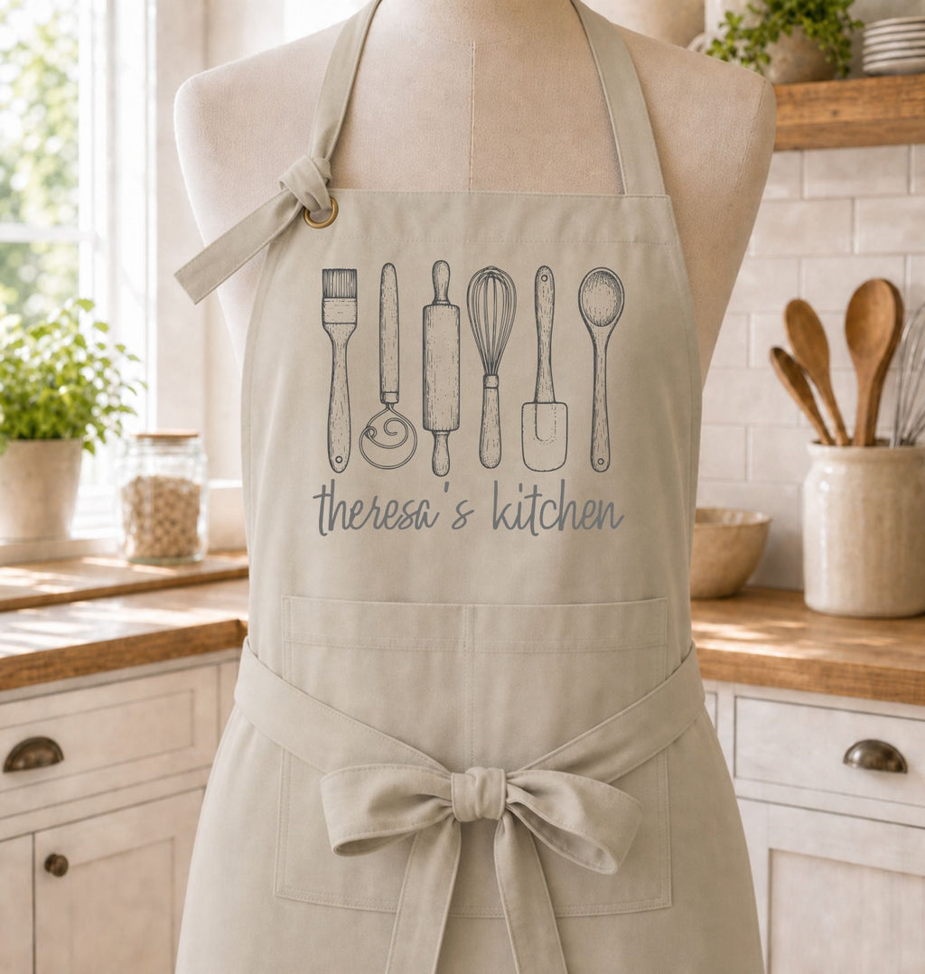 A beige kitchen apron displayed on a dress form in a bright, real kitchen. The apron features a gray line art graphic of various kitchen utensils and the text 'theresa's kitchen' printed on the chest.